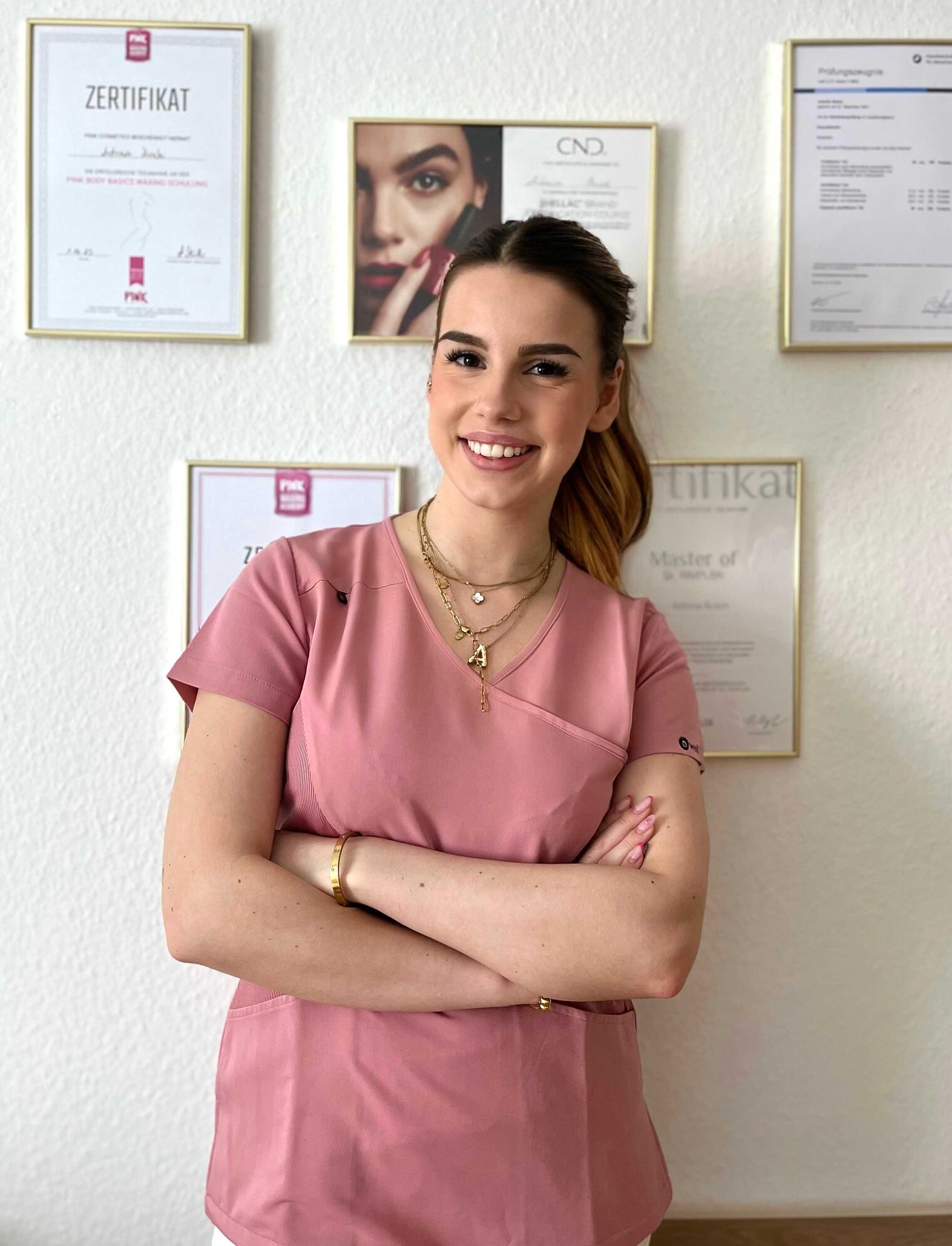 Frau steht vor Wand mit Zertifikaten in Rahmen, verschränkte Arme, lächelt